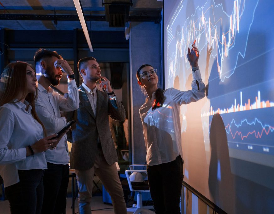 Standing together. Male leader talking to employees, showing the plan on the projector in office of stock exchange company.